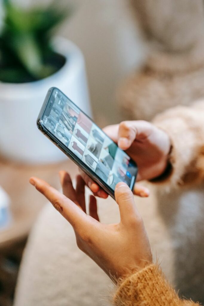 Crop unrecognizable female demonstrating photos of clothes on mobile phone screen at home on blurred background